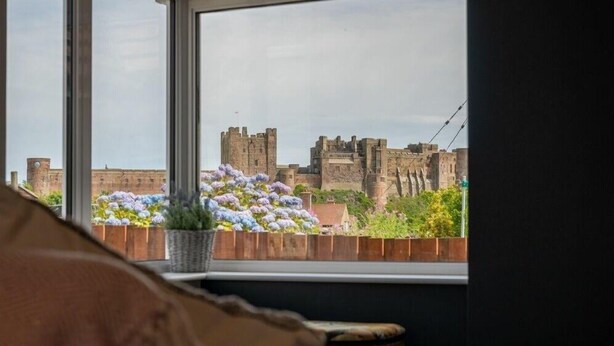 A view of a historic castle through a window with a wooden ledge and potted flowers. - Castle View (Bamburgh)
