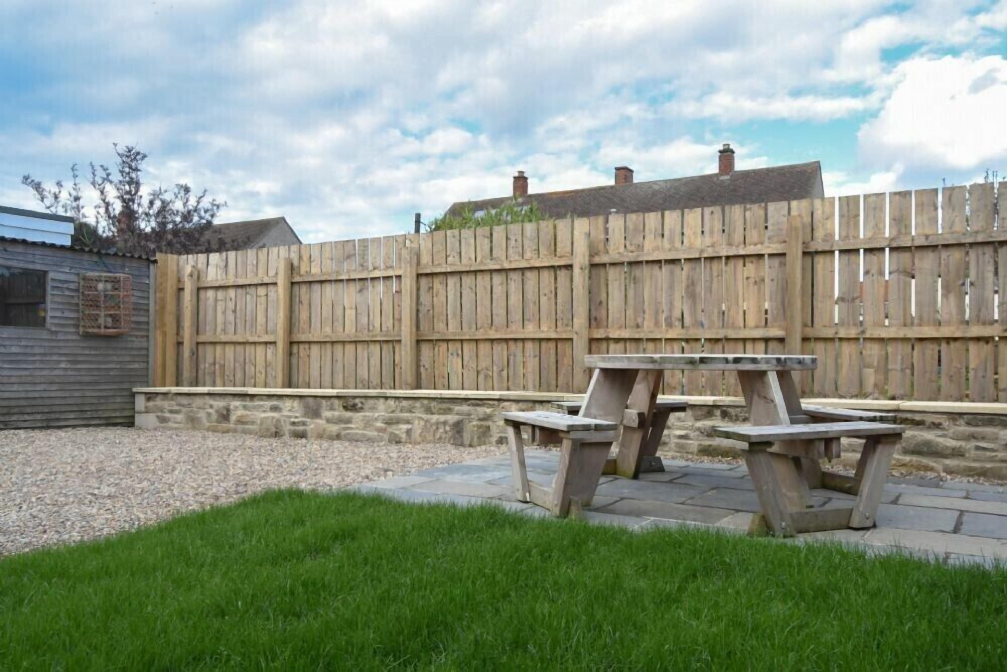 A backyard with a wooden fence, a stone wall, and two picnic tables.