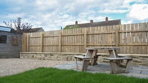 Un giardino recintato da una staccionata di legno, con un muro in pietra e due tavoli da picnic.