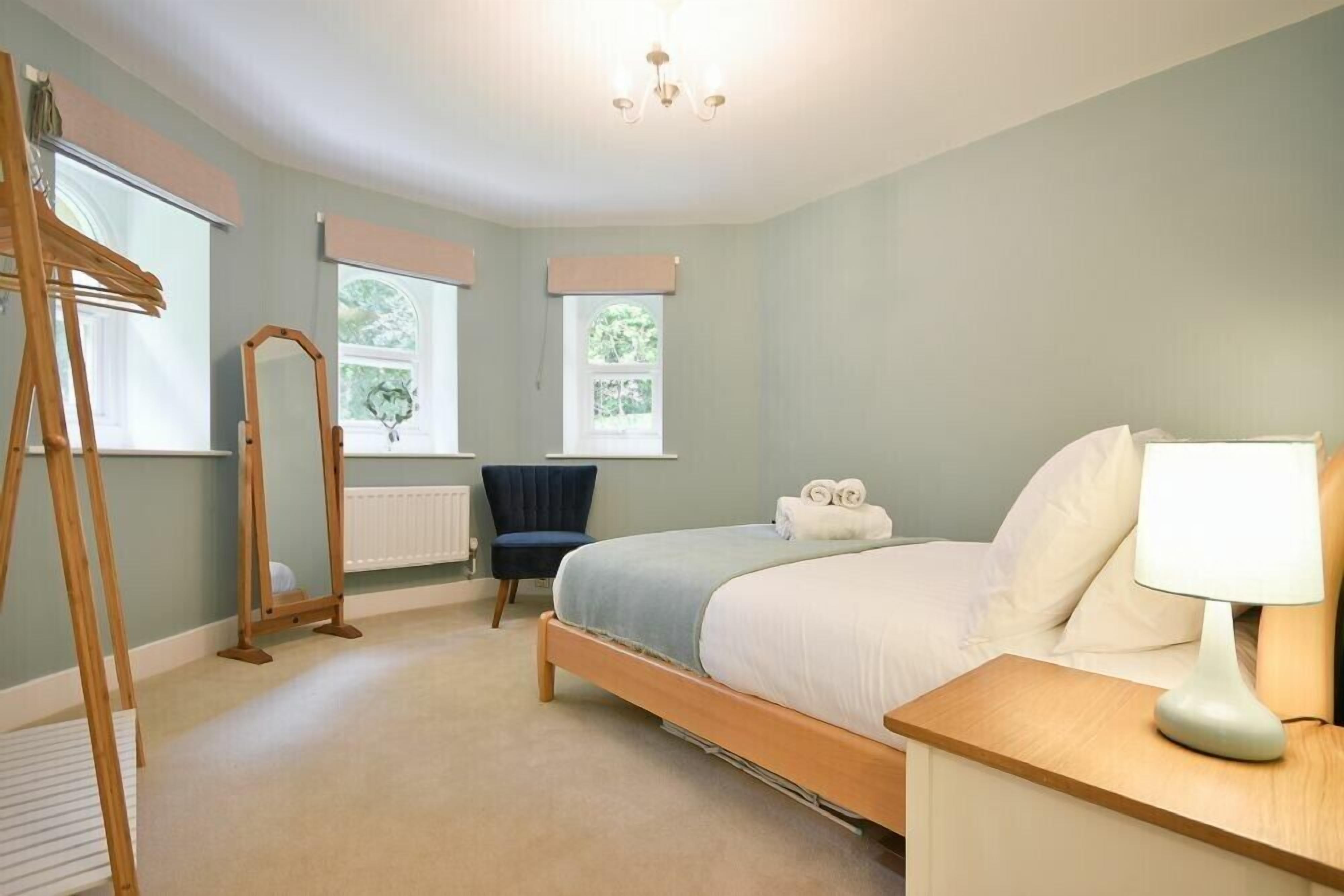 A bedroom with a bed, bedside table, lamp, chair, windows, radiator, and a ladder.