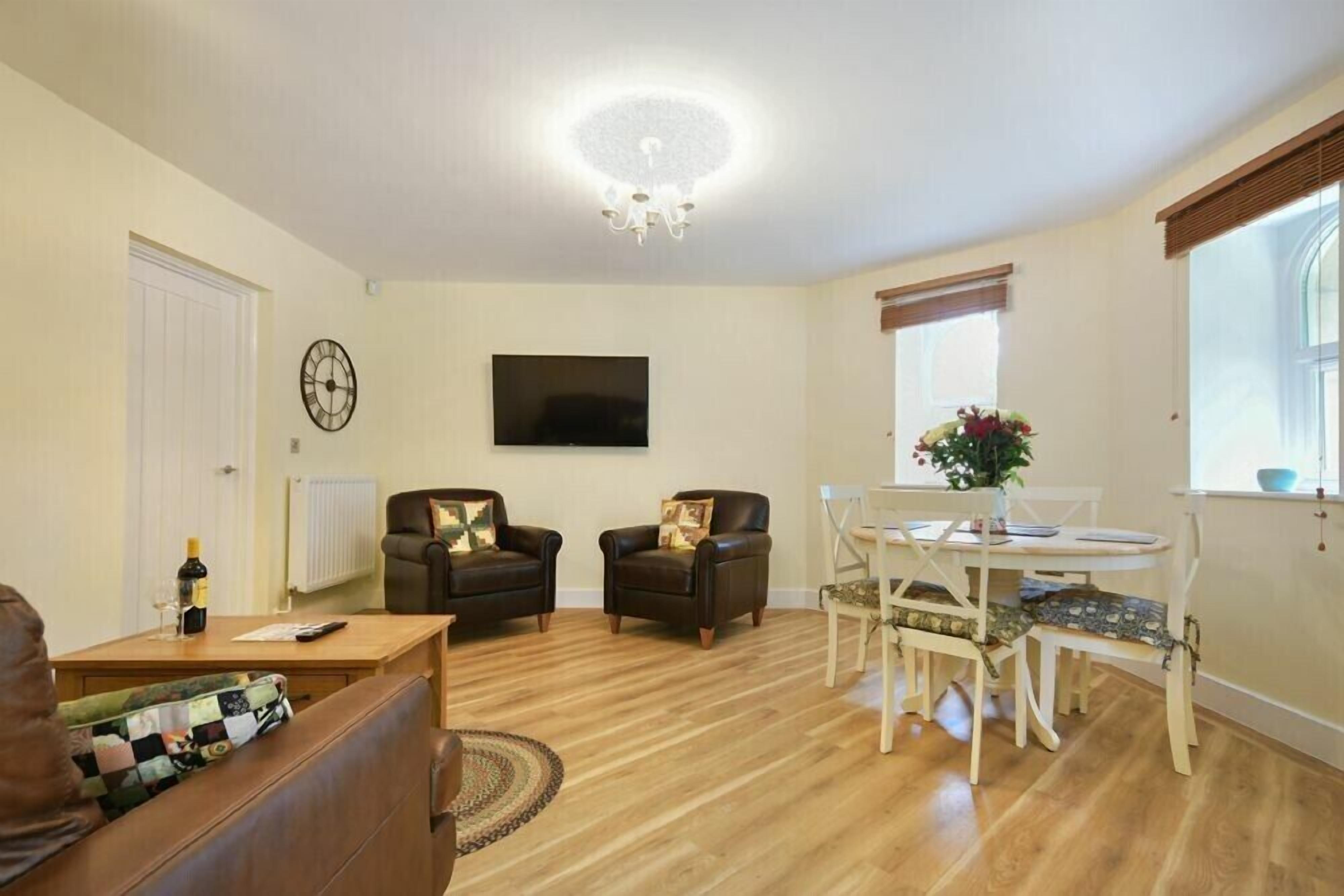 A living room with a wooden floor, two leather armchairs, a white dining table with chairs, a TV mounted on the wall, and a chandelier.