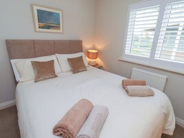 A neatly made bed with pillows and two rolled towels, a bedside lamp, and a window with blinds.