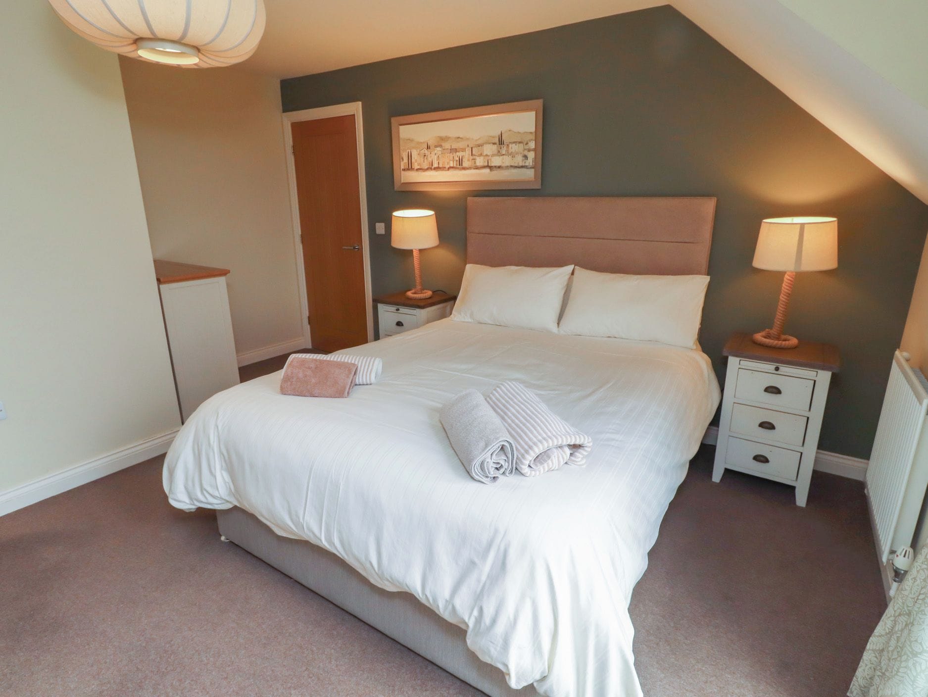 A bedroom with a bed, two bedside tables, a nightstand, a door, and a wall-mounted lamp.