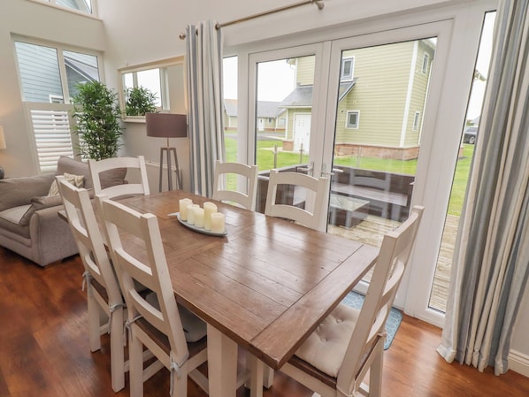 A dining area with a wooden table, white chairs, and a view through glass doors to a backyard. - The Beach Hut (Chathill)