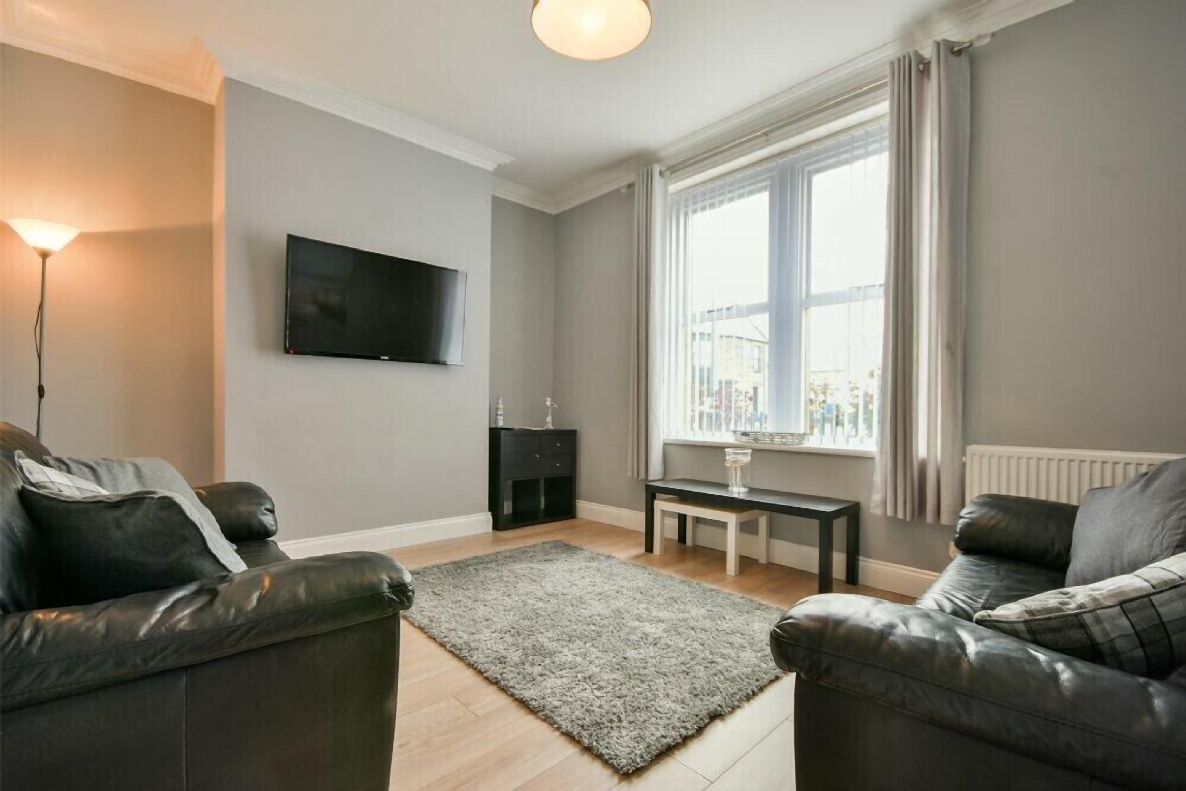 Un salon moderne avec deux canapés en cuir, un tapis à poils longs de couleur grise, un téléviseur à écran plat fixé au mur, une petite table noire et une fenêtre avec des rideaux.