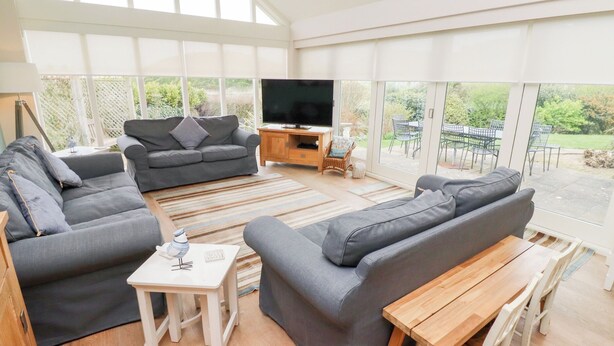 Ein großzügiger Wohnbereich mit großem Fernseher, grauen Sofas, einem Holztisch und Blick auf den Garten durch große Fenster.