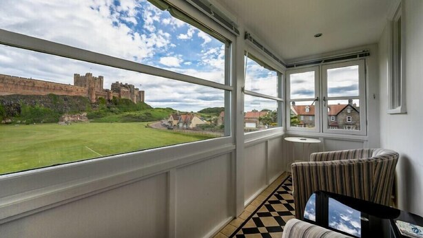 Ein Balkon mit großen Fenstern, Blick auf ein Schloss und ein grünes Feld. Zwei gestreifte Sessel und ein kleiner Tisch mit einem Bilderrahmen sind vorhanden.