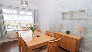 A dining area with a wooden table and chairs, a vase with white flowers, a small cabinet with lamps, and a window with curtains. - Bay View (Morpeth)