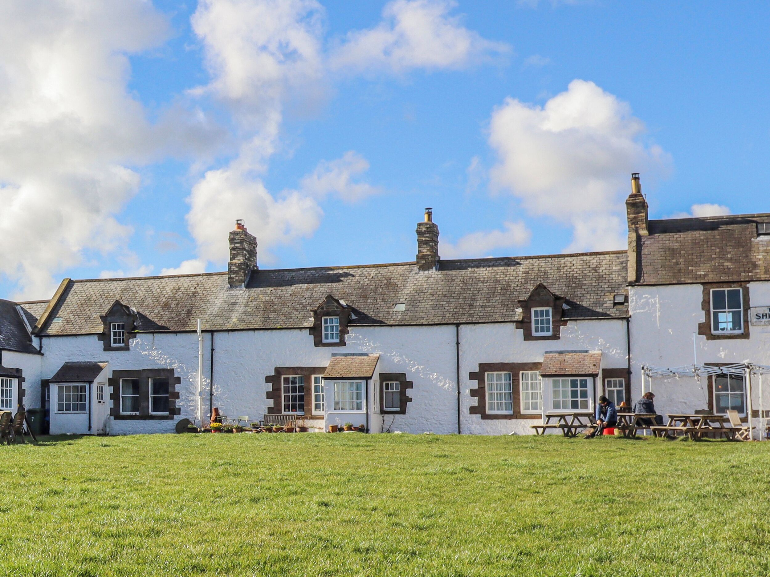 A row of white buildings with multiple chimneys, a grassy field in the foreground, and a clear blue sky with clouds.
