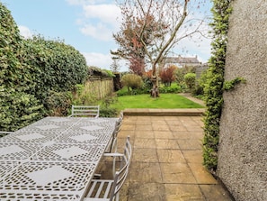 A patio with a metal table and chairs, surrounded by greenery and a stone pathway leading to a house. - St Marys Cottages No4 (Alnwick)