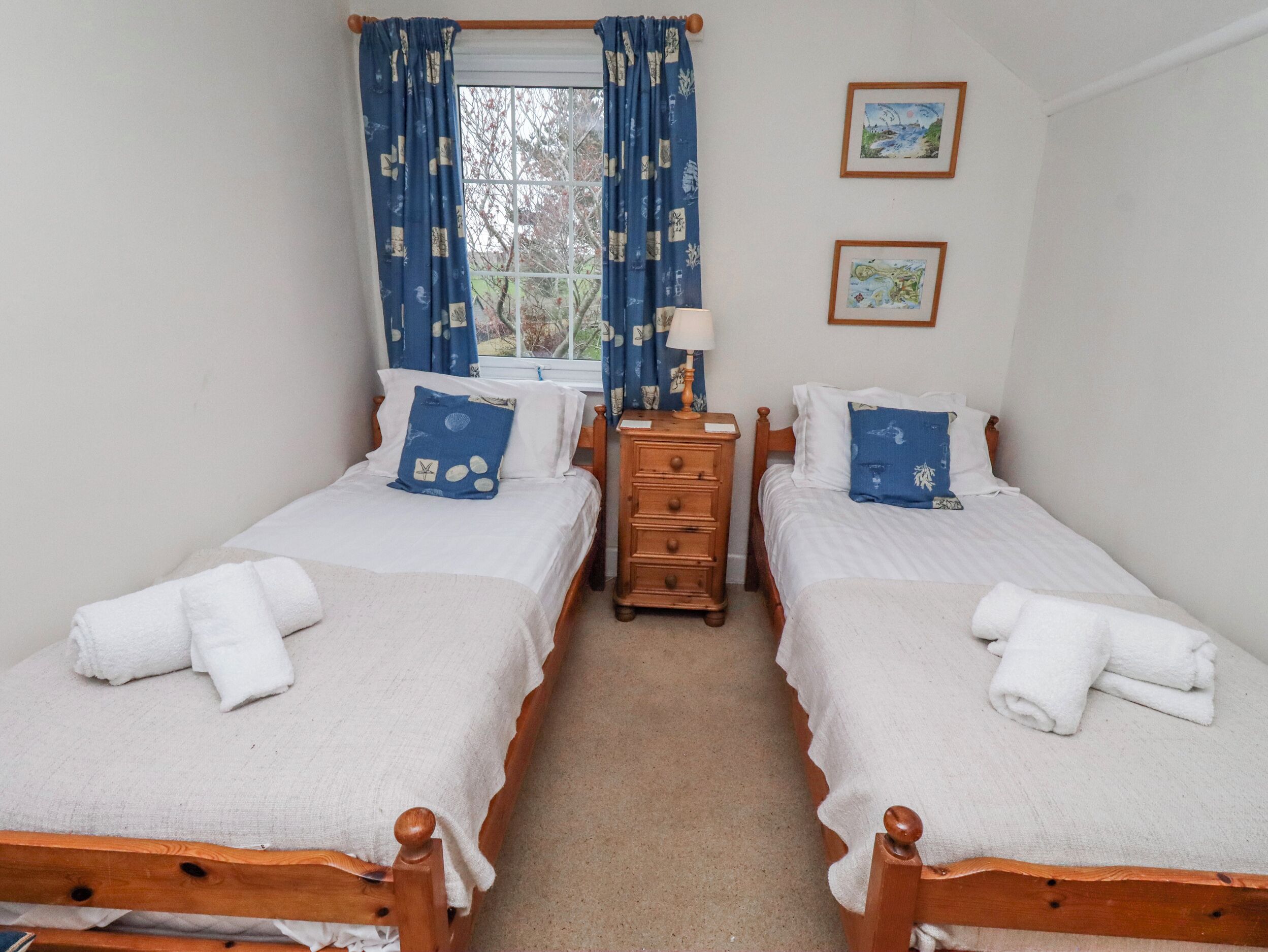 A room with two single beds, a wooden dresser, a window with curtains, and two framed pictures on the wall.