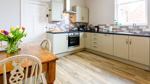A modern kitchen with white cabinets, a wooden dining table, and a window with curtains. - Baysyde (Alnwick)