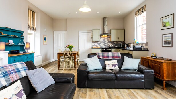 A modern living room with a black leather sofa, a wooden dining table, and a kitchen in the background. - Baysyde (Alnwick)