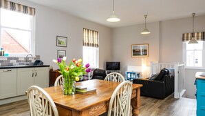 A dining area with a wooden table, white chairs, and a vase of flowers. There is a kitchen in the background with white cabinets and a window. - Baysyde (Alnwick)