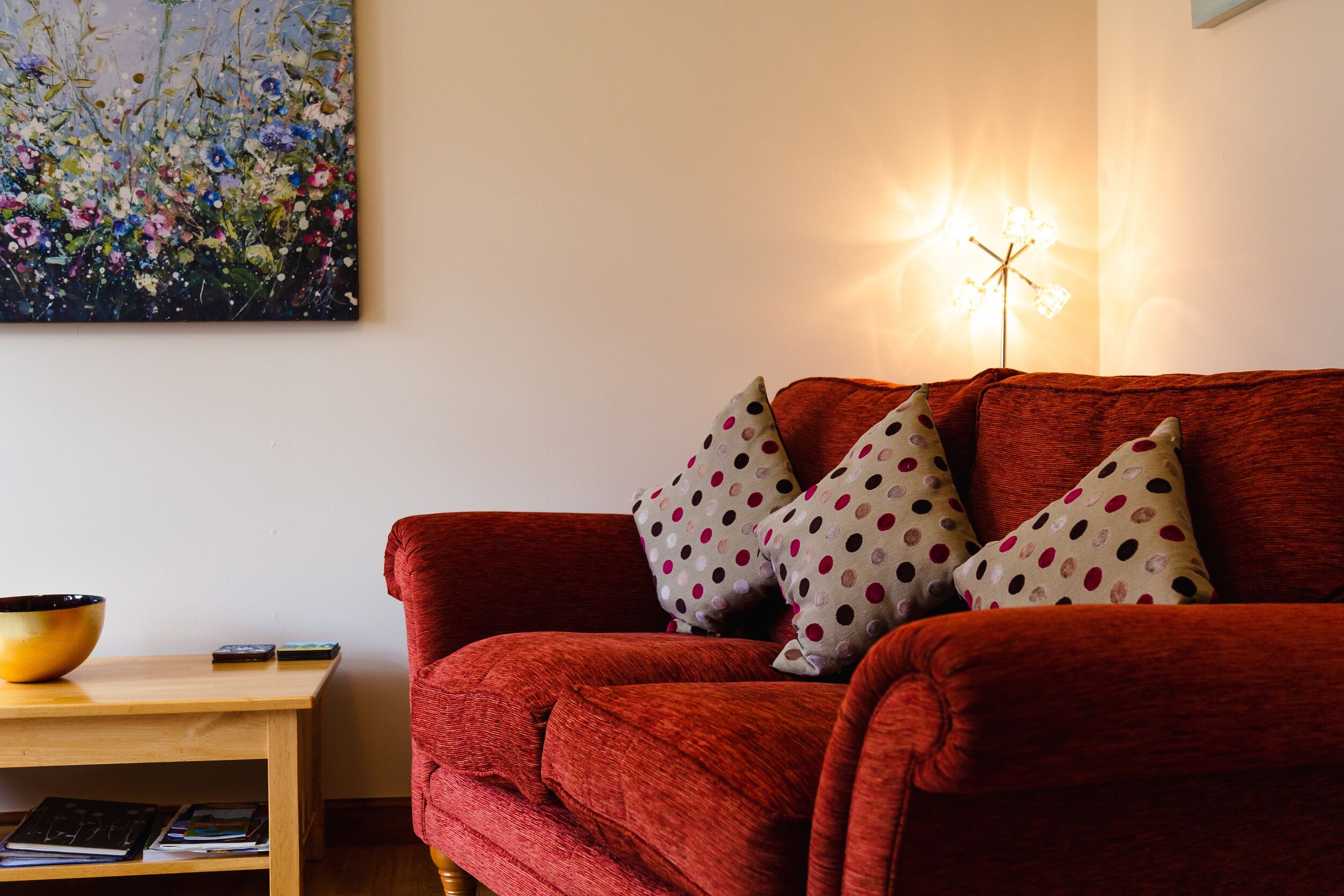 A red sofa with polka dot cushions, a wooden side table with a bowl and books, and a wall-mounted light fixture.