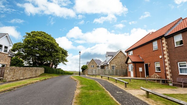 A residential street with houses, a paved road, and a grassy area. - The Apartment (Bamburgh)