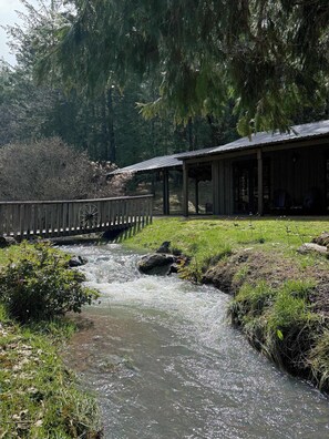 A riverside walkway with a wooden bridge, a covered pavilion, and a grassy area.