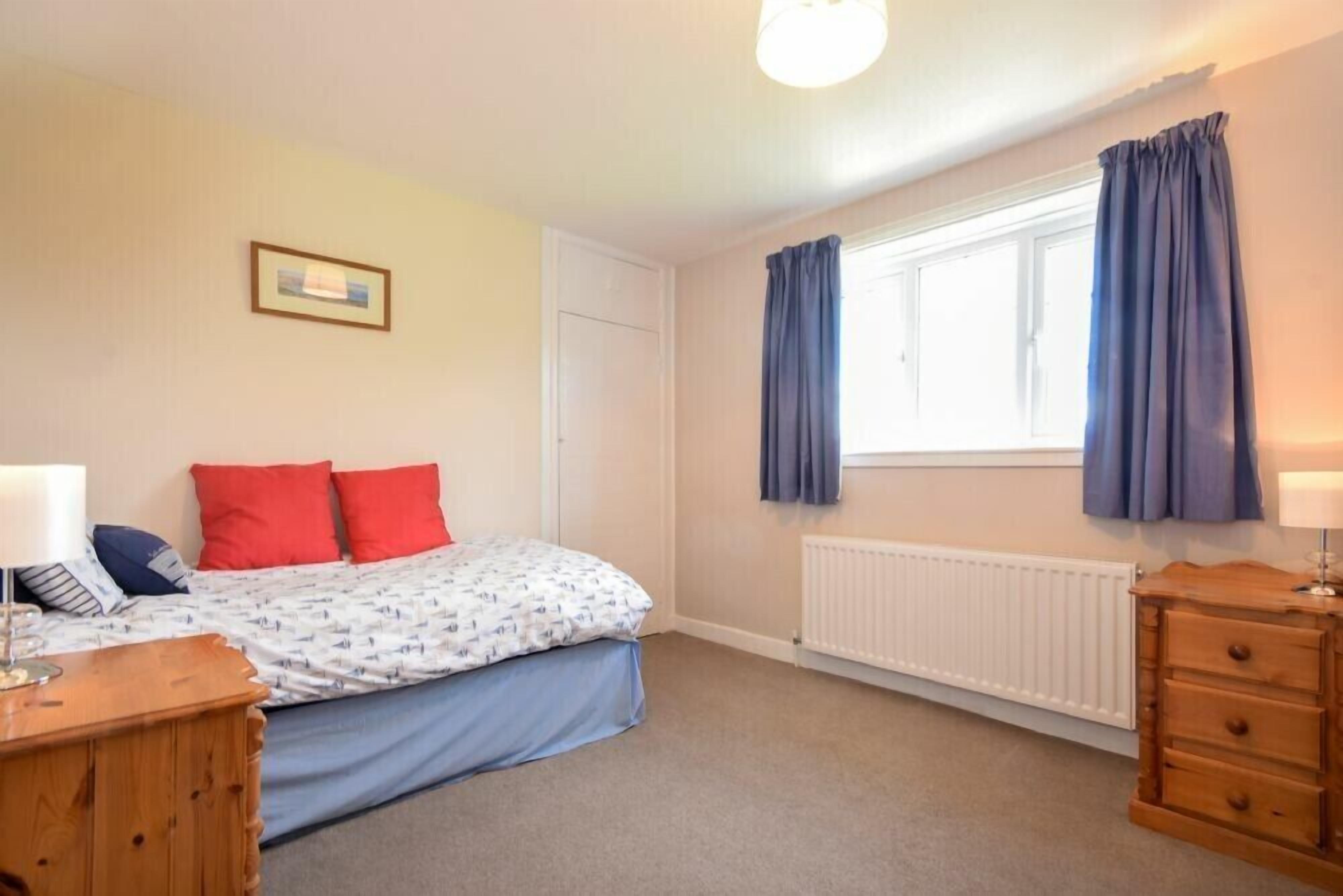 A bedroom with a bed, a dresser, a nightstand, a window with curtains, a radiator, and a framed picture on the wall.