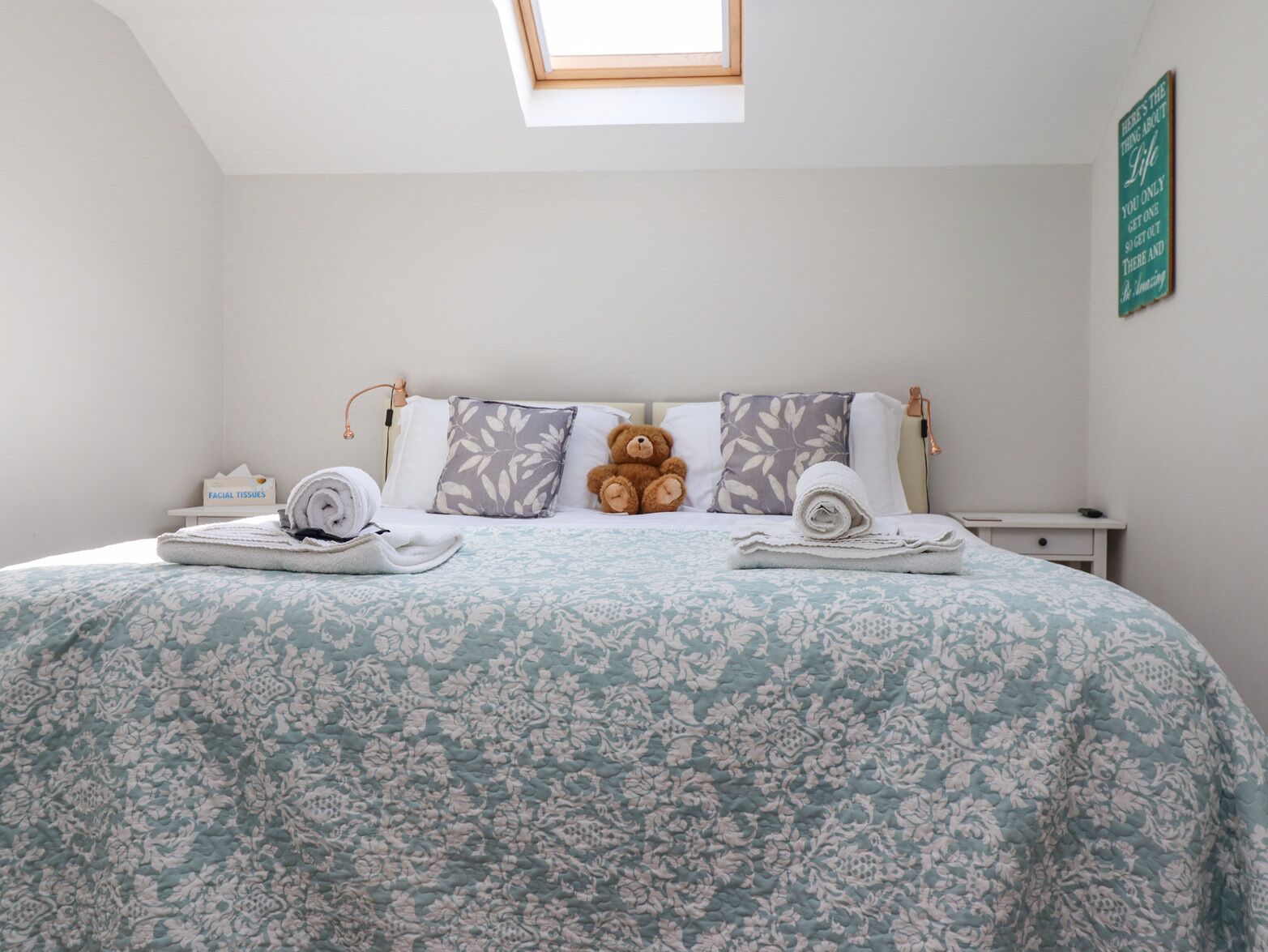 A bedroom with a skylight, a bed with floral bedding, two pillows, a teddy bear, and a bedside table with a lamp.