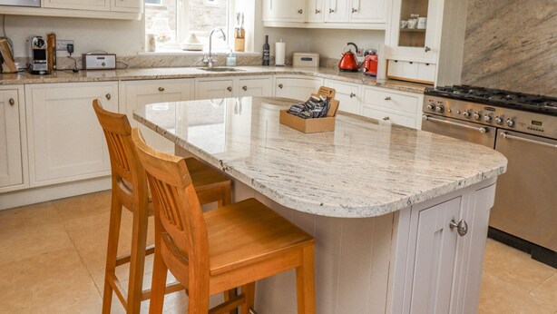 A kitchen with white cabinets, a marble countertop, a central island with stools, a stove, and a window with red floral curtains. - The Mustard Pot (Morpeth)