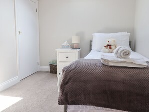 A neatly made bed with a brown blanket, a white pillow, and a teddy bear. There is a nightstand with a lamp, a box of tissues, and a trash can. - The Mustard Pot (Morpeth)
