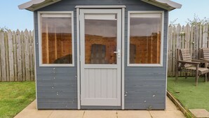 A small, single-story shed with a grey exterior and a white door. It has two large windows on the front side, and a wooden fence is visible in the background. - The Mustard Pot (Morpeth)