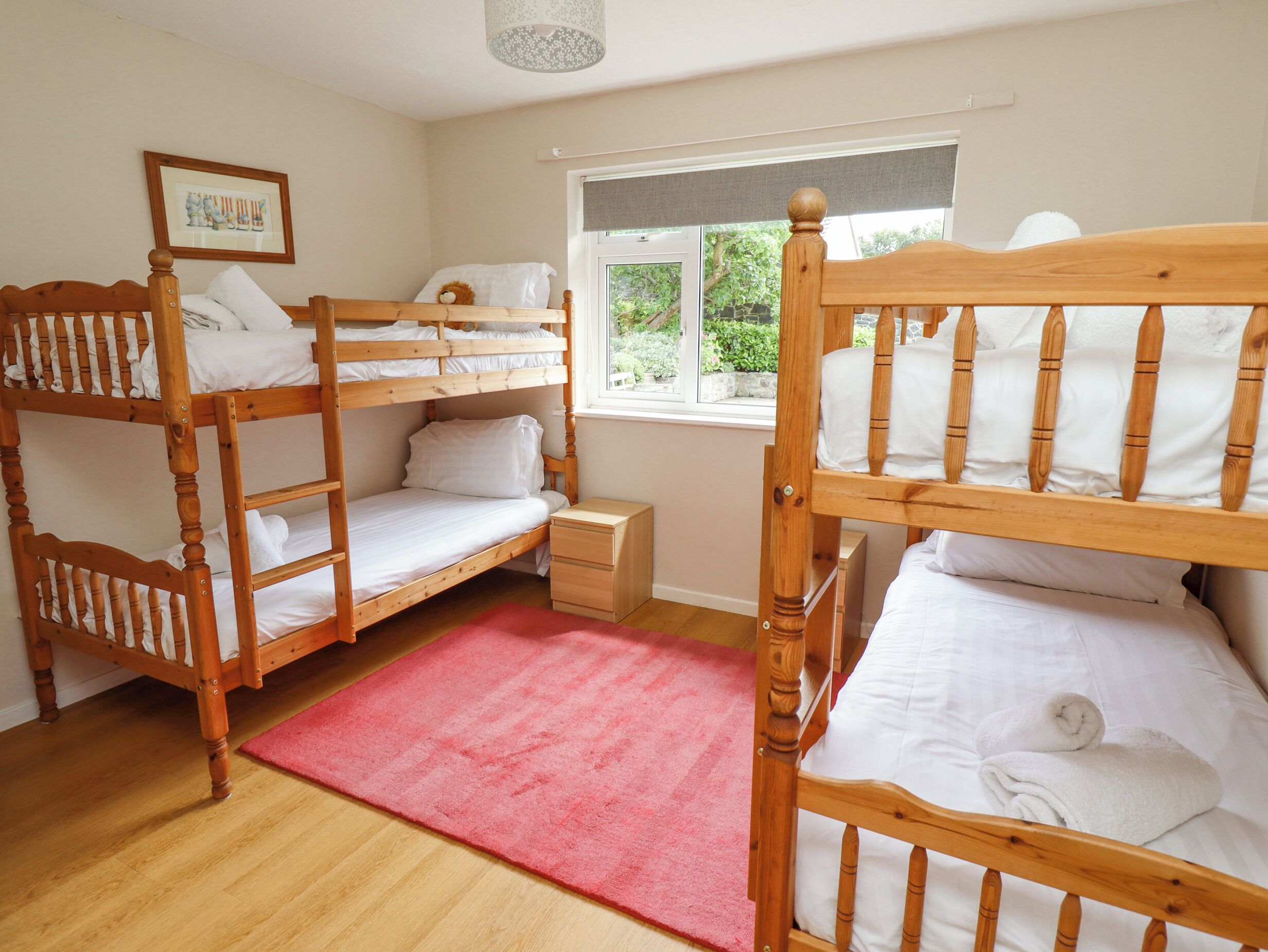 A room with bunk beds, a small bedside table, a window with a view of greenery, and a framed picture on the wall.