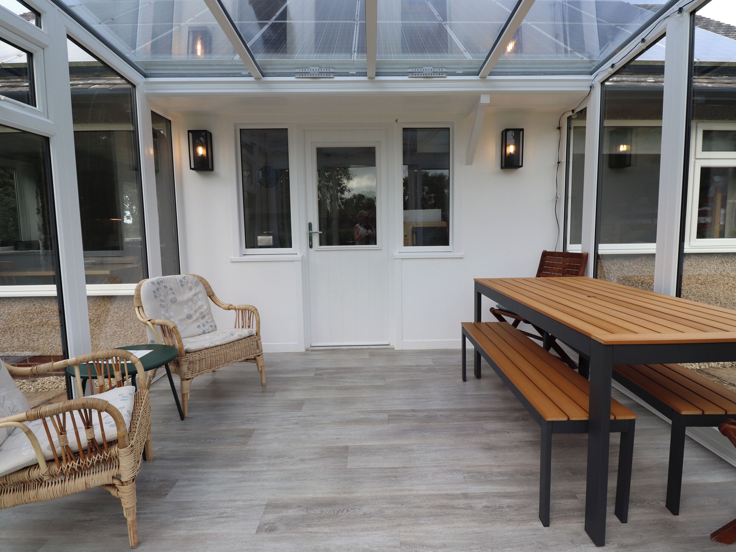 A covered patio area with a wooden table and benches, wicker chairs, and a glass ceiling.