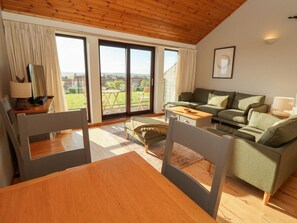 A cozy living room with a wooden ceiling, a green sofa, a wooden dining table, and a view through large glass doors.