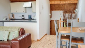 A cozy living room with a brown leather sofa, a wooden dining table with chairs, and a kitchen area with white cabinets and a kettle.