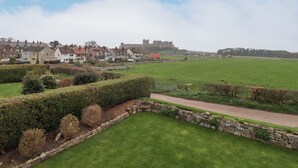 A rural landscape with a castle in the background, a green field, and a row of houses. - Whitestacks (Bamburgh)