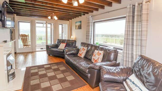 A spacious living room with a large leather sofa, a wooden floor, and a view of the outdoors through a large window. - Whitestacks (Bamburgh)