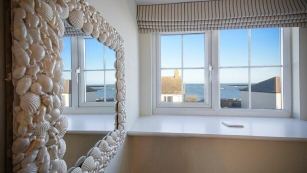 A room with a large mirror featuring a shell design, a window with a view of the sea and buildings, and a white bench. - Coast View (Chathill)