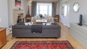 A spacious living room with a grey sofa, a coffee table, and a red patterned rug. There is a fireplace with a mantel and a painting on the wall. - The Lookout (Alnwick)