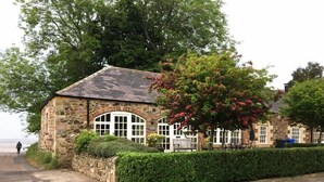 A stone building with a sloped roof, large windows, and a hedge in front. - Lapwing Cottage (Bamburgh)