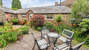 A stone building with a garden patio featuring wicker furniture and a variety of plants. - Nurse's Cottage (Alnwick)