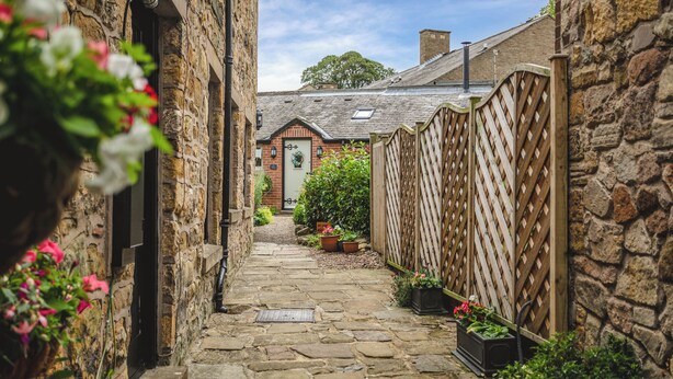 A narrow stone pathway leading to a brick building with a green door, surrounded by wooden fences and potted plants. - Nurse's Cottage (Alnwick)