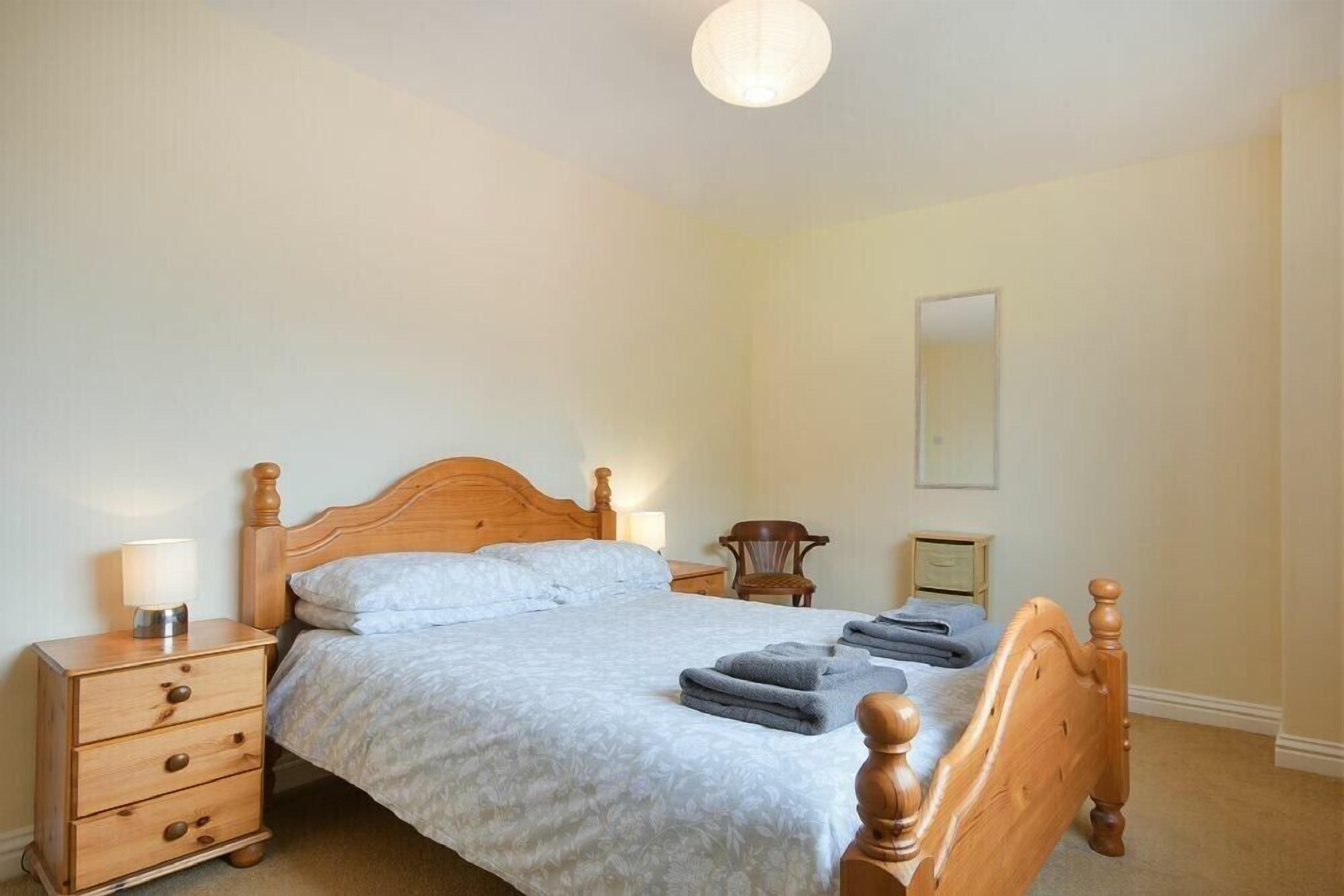 A bedroom with a wooden bed, a nightstand with a lamp, a chair, and a small bedside table.