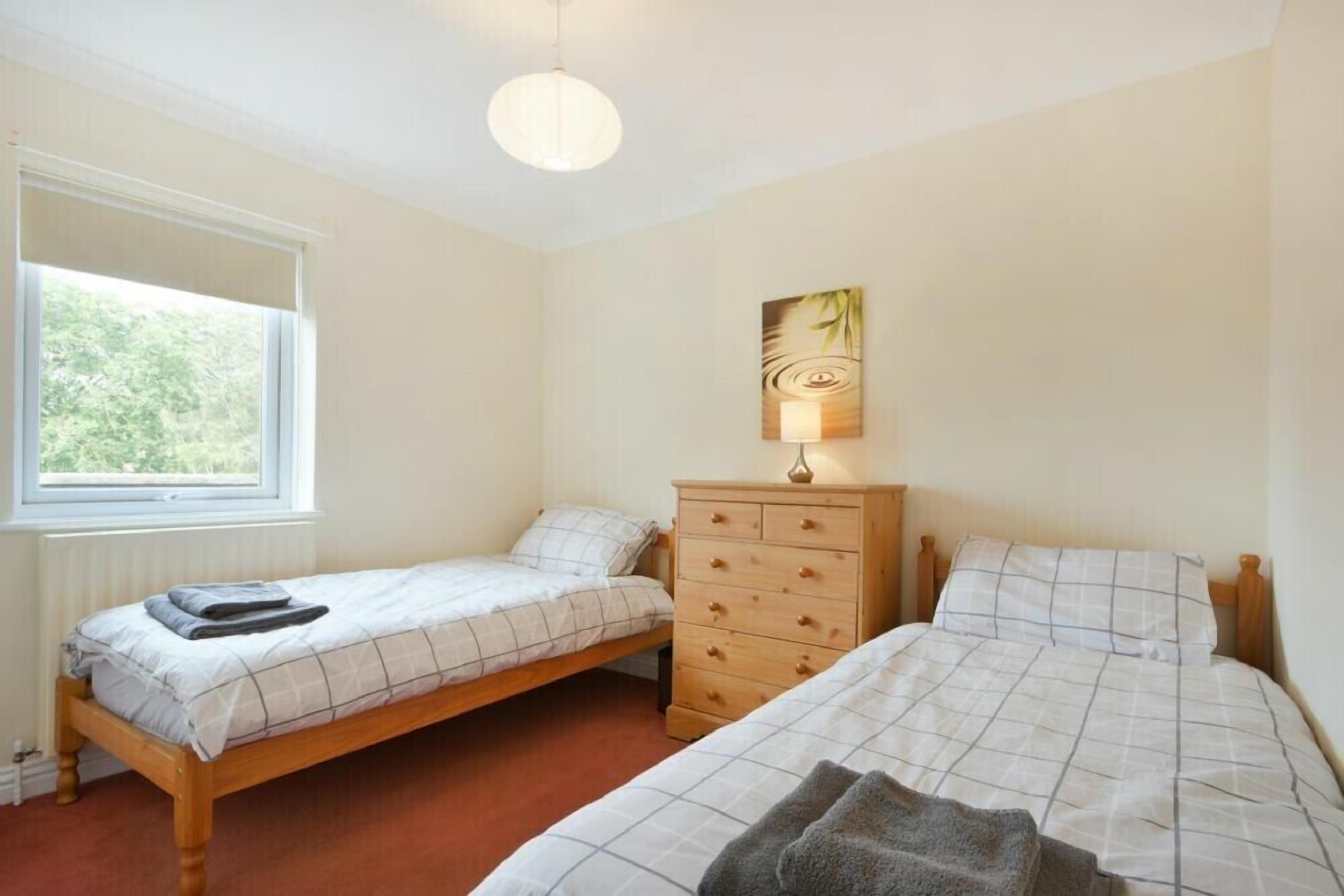Une chambre avec deux lits simples, une commode en bois, une lampe de chevet et une fenêtre laissant entrer la lumière naturelle.