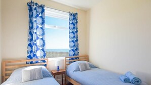 Ein Zimmer mit zwei Einzelbetten, einem Holz-Kopfteil, einem kleinen Nachttisch mit Lampe und einem Fenster mit blumigen, blauen Vorhängen.