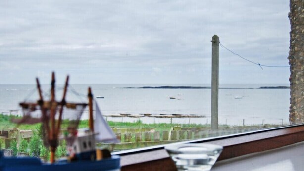 Blick aus einem Fenster mit einem Modellschiff und einem Glas Wasser auf einem Tisch, mit Blick auf ein ruhiges Meer und einen Leuchtturm.