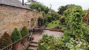 A stone building with a chimney, a patio with chairs, and a garden with various plants and a lawn. - The Book House (Morpeth)