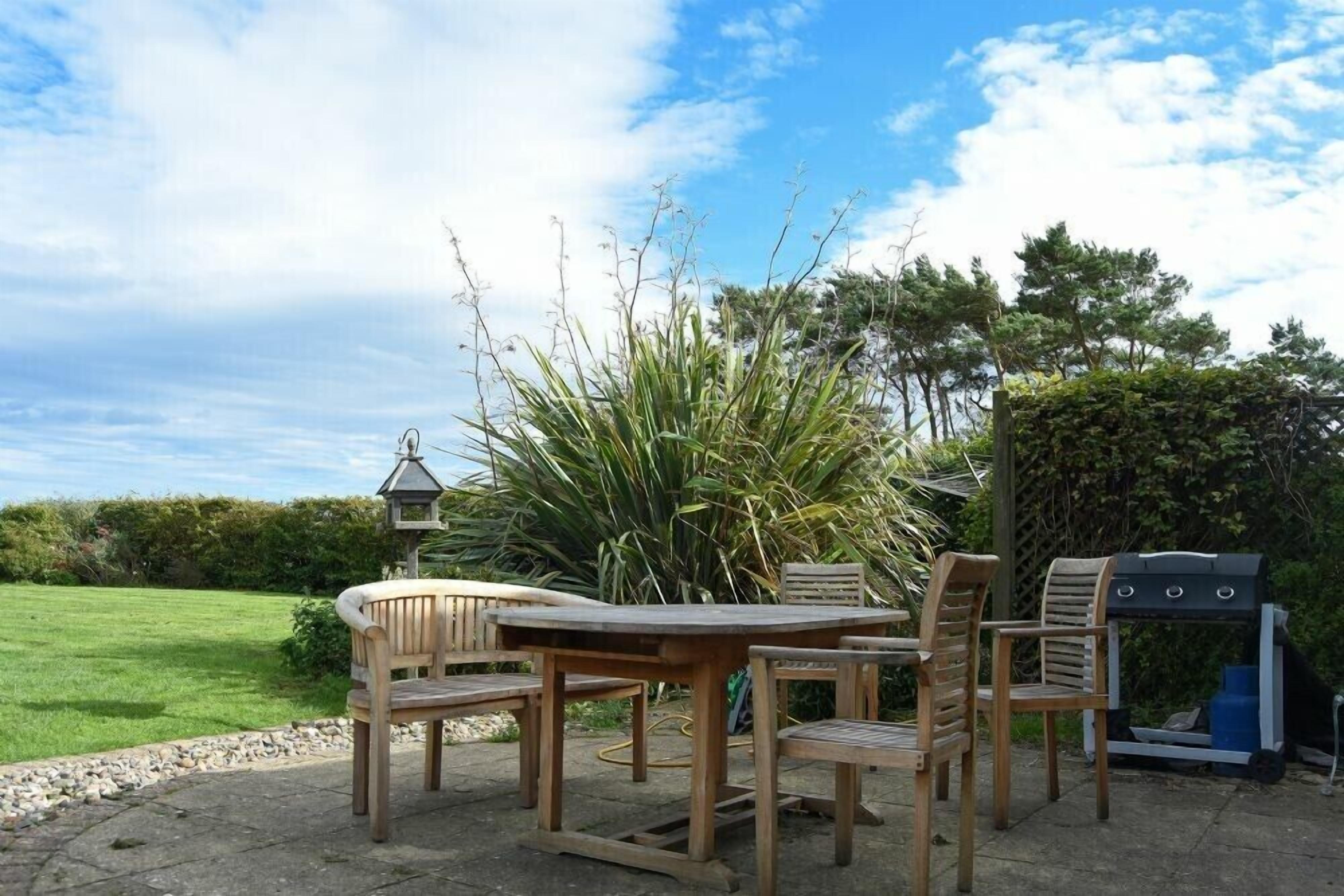 A patio with a wooden table and chairs, a barbecue grill, and a garden with tall plants and a hedge.