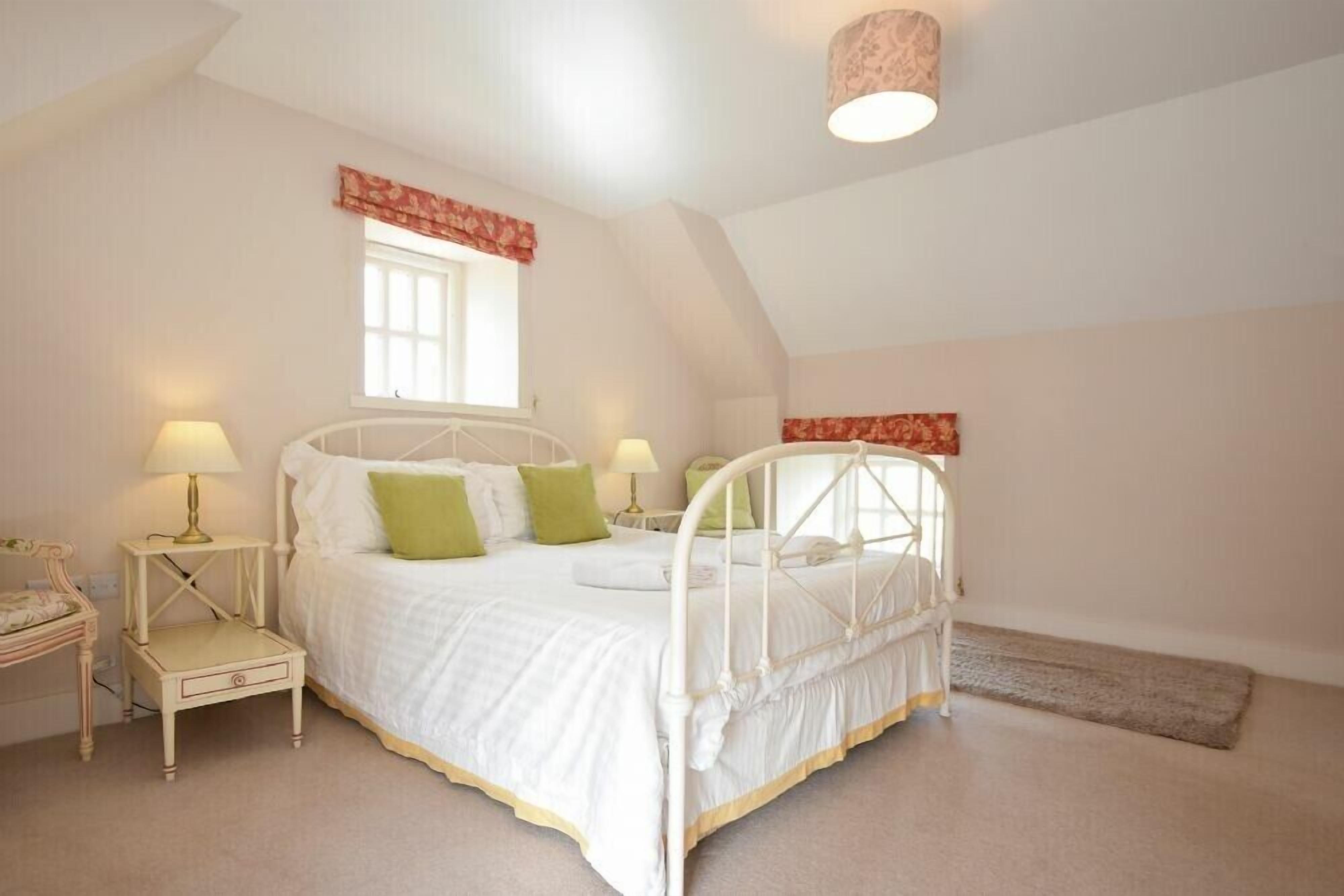 A bedroom with a white bed, green pillows, a small bedside table with a lamp, and a window with a red valance.