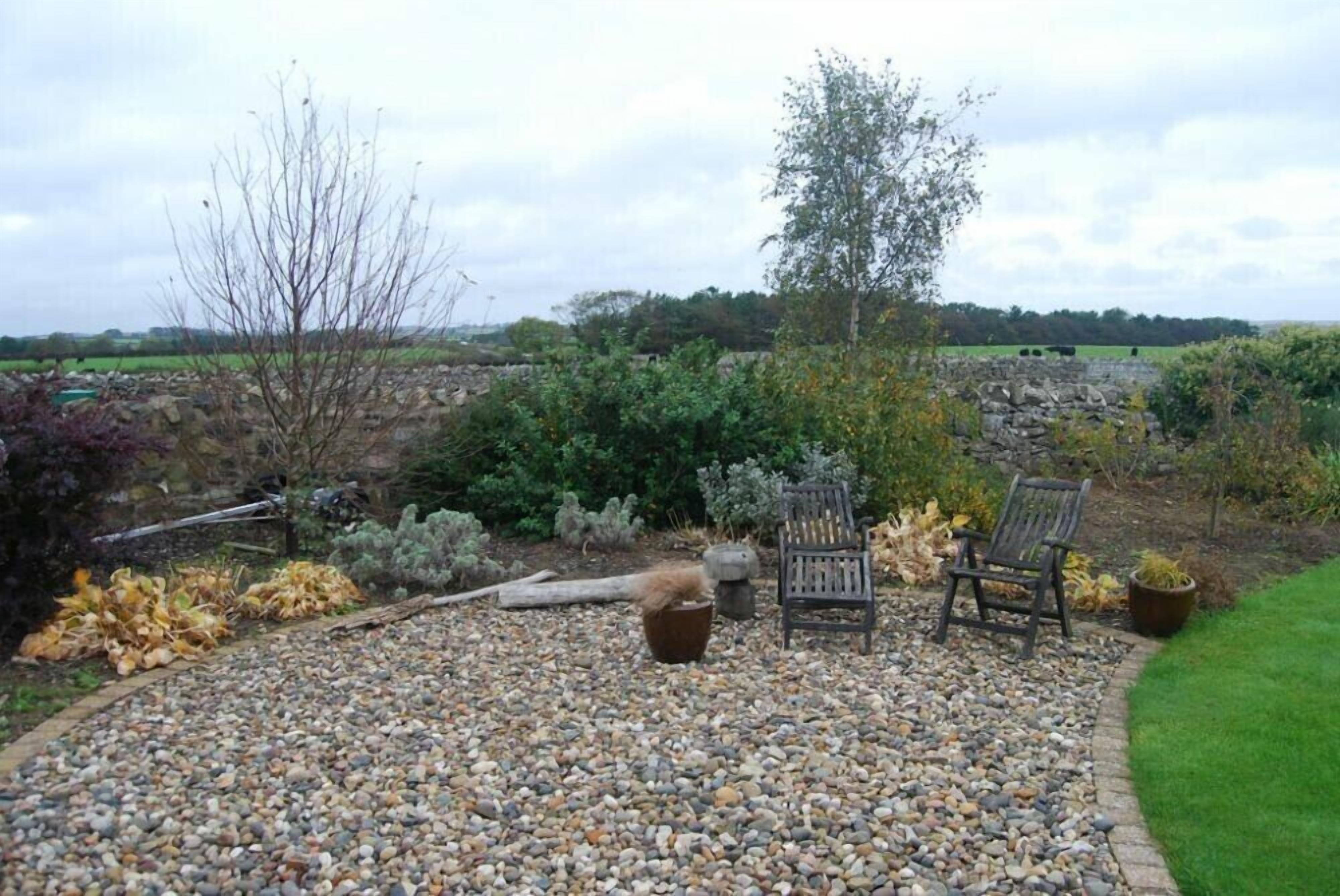A garden patio with a gravel path, two wooden chairs, and a potted plant.