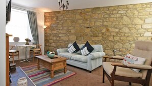 Ein gemütliches Wohnzimmer mit Steinwand, einem hellen Sofa mit Streifen-Kissen, einem Holztisch und einem beigefarbenen Sessel, sowie einem bunten Streifen Teppich.