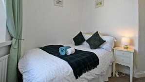 Ein ordentlich gemachtes Bett mit weißer Daunendecke, einer grün-schwarz karierten Decke und zwei aufgerollten Handtüchern. Ein kleiner Nachttisch mit Lampe und Schublade ist ebenfalls vorhanden.
