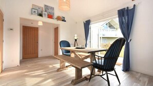 Un espace repas avec une table et des bancs en bois, une chaise bleue, un luminaire suspendu et une vue sur un balcon.
