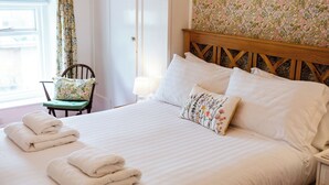 Ein ordentlich gemachtes Bett mit weißen Leinentüchern und floralen Kissen, ein Sessel mit einem floralen Sitzkissen sowie ein Fenster mit einem floralen Vorhang.