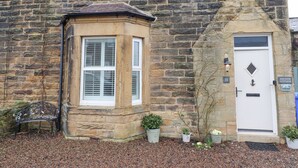 Un bâtiment en pierre avec une fenêtre d’angle, une porte portant une plaque avec un numéro, et des plantes en pot.